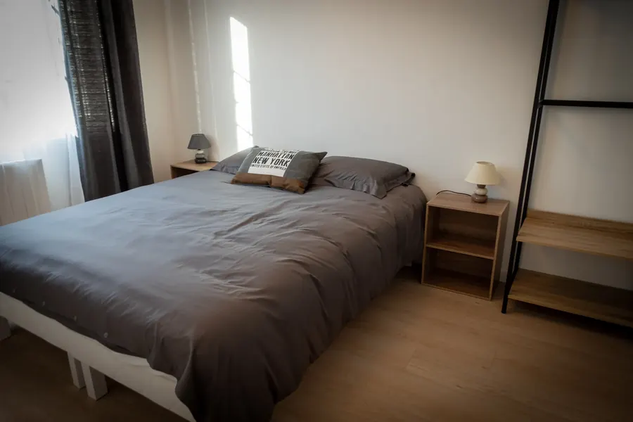 Le lit fait, linge gris, une lampe de chevet et une étagère en bois