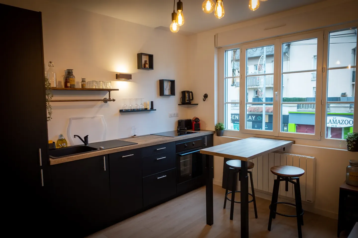 La cuisine noire, la table haute et ses deux tabourets, la fenêtre sur l'avenue